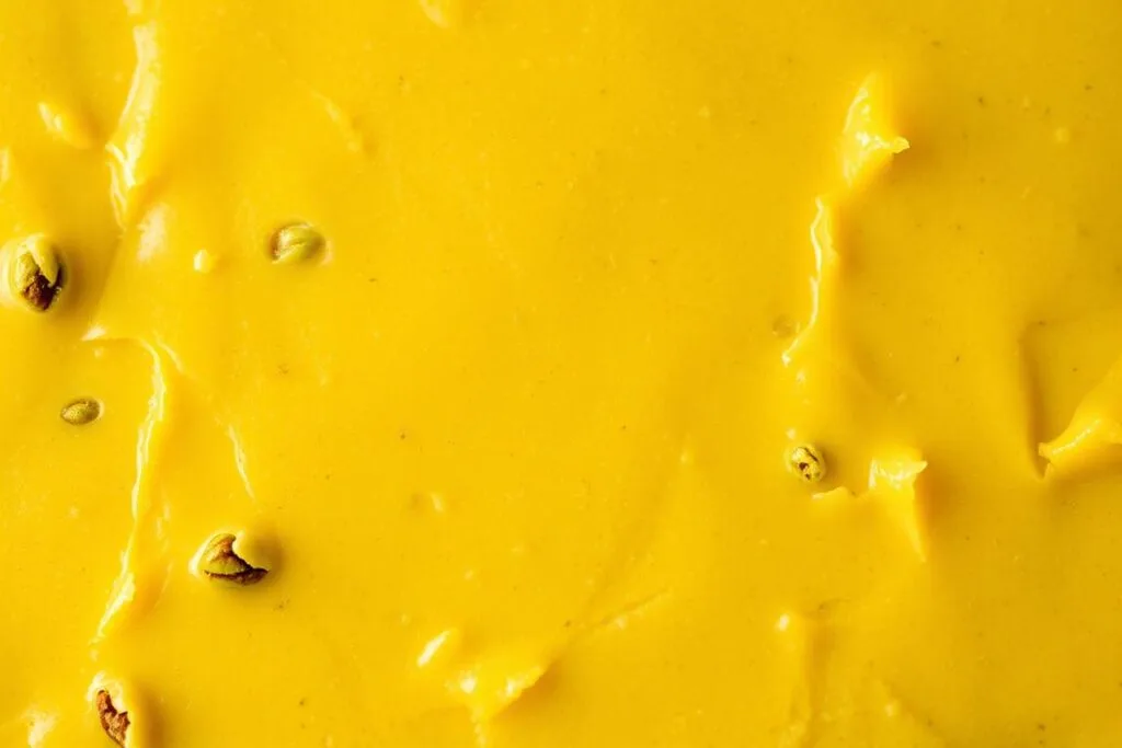 A closeup view of freshly made pistachio fudge, showcasing its luscious texture and rich color. The surface is smooth and glossy, with a slight sheen that catches the light. Tiny flecks of vibrant green pistachio kernels are visible throughout the creamy, indulgent confection. Warm lighting from above casts soft shadows, emphasizing the depth and luxurious mouthfeel of the fudge. The composition is tightly framed, filling the frame and drawing the viewer's attention to the decadent texture. A sense of artisanal craft and attention to detail is evident in every aspect of the image.