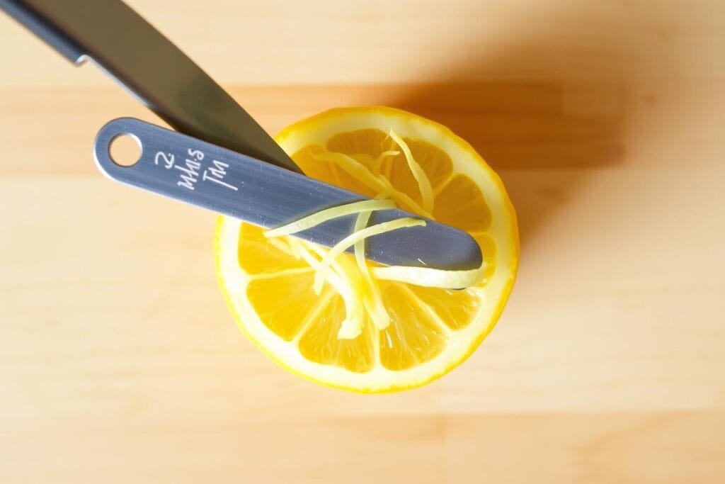 Lemon zesting in progress, with a sharp zester tool gliding across the vibrant yellow rind of a fresh lemon, creating thin, curled strips of aromatic zest. Against a light-toned wooden surface, the action is captured from an overhead perspective, highlighting the intricate details of the zest extraction technique. Subtle natural lighting casts soft shadows, emphasizing the texture and juiciness of the lemon. The image conveys a sense of culinary expertise and the preparation for a zesty, flavorful lemon loaf. Lemon zesting in progress, with a sharp zester tool gliding across the vibrant yellow rind of a fresh lemon, creating thin, curled strips of aromatic zest. Against a light-toned wooden surface, the action is captured from an overhead perspective, highlighting the intricate details of the zest extraction technique. Subtle natural lighting casts soft shadows, emphasizing the texture and juiciness of the lemon. The image conveys a sense of culinary expertise and the preparation for a zesty, flavorful lemon loaf.