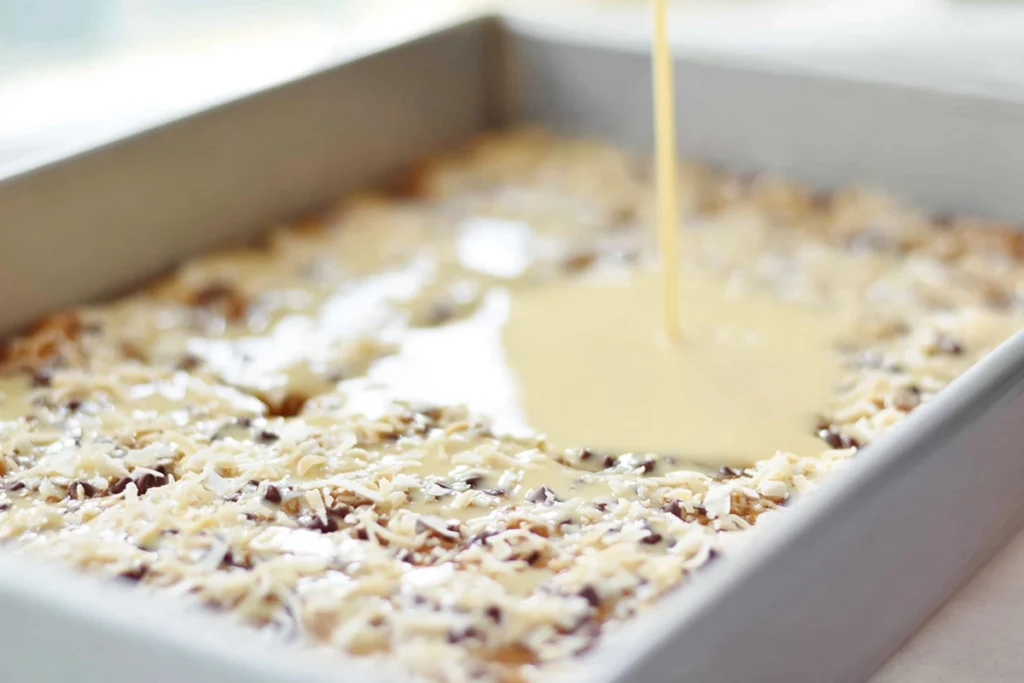 close-up of sweetened condensed milk pouring over graham crust for magic cookie bars